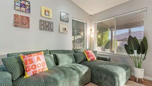A green sectional sofa with two pillows, a potted plant, and framed pictures on the wall. - Vibrant Home – Heated Pool, Arcade, & Fire pit (Phoenix)