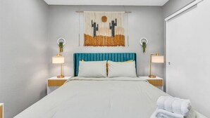 A modern bedroom with a bed, two bedside tables, a wall-mounted artwork, and a door. - Vibrant Home – Heated Pool, Arcade, & Fire pit (Phoenix)