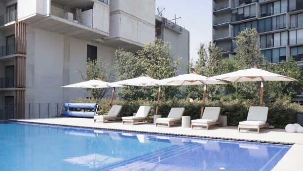 A swimming pool with lounge chairs and umbrellas, surrounded by greenery and modern buildings. - City-view condo w/ pool & free parking (Guadalajara)