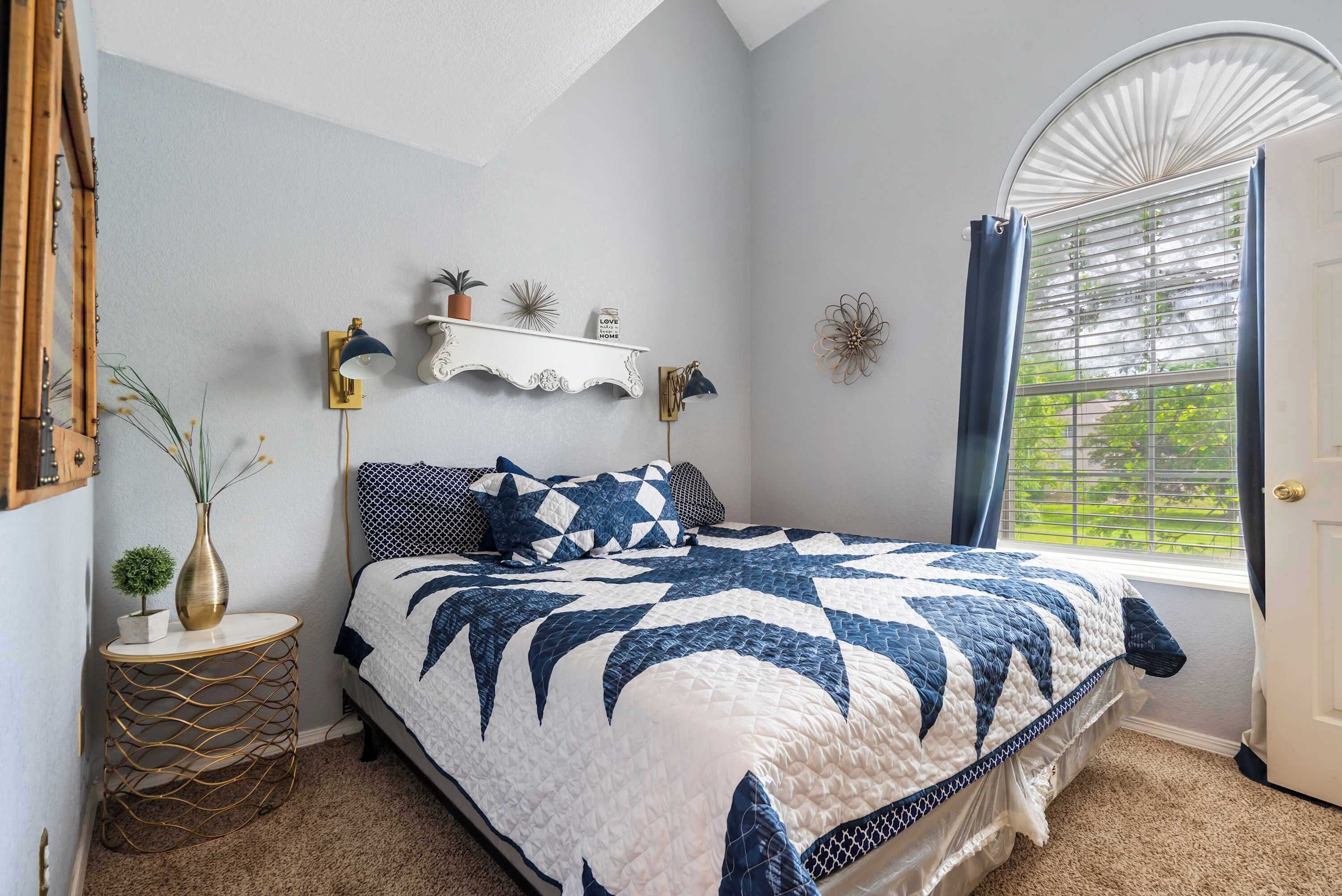 Ein Schlafzimmer mit einem Bett, einem Nachttisch, einer kleinen Pflanze, einem an der Wand befestigten Regal mit Dekoration und einem Fenster mit Vorhängen.