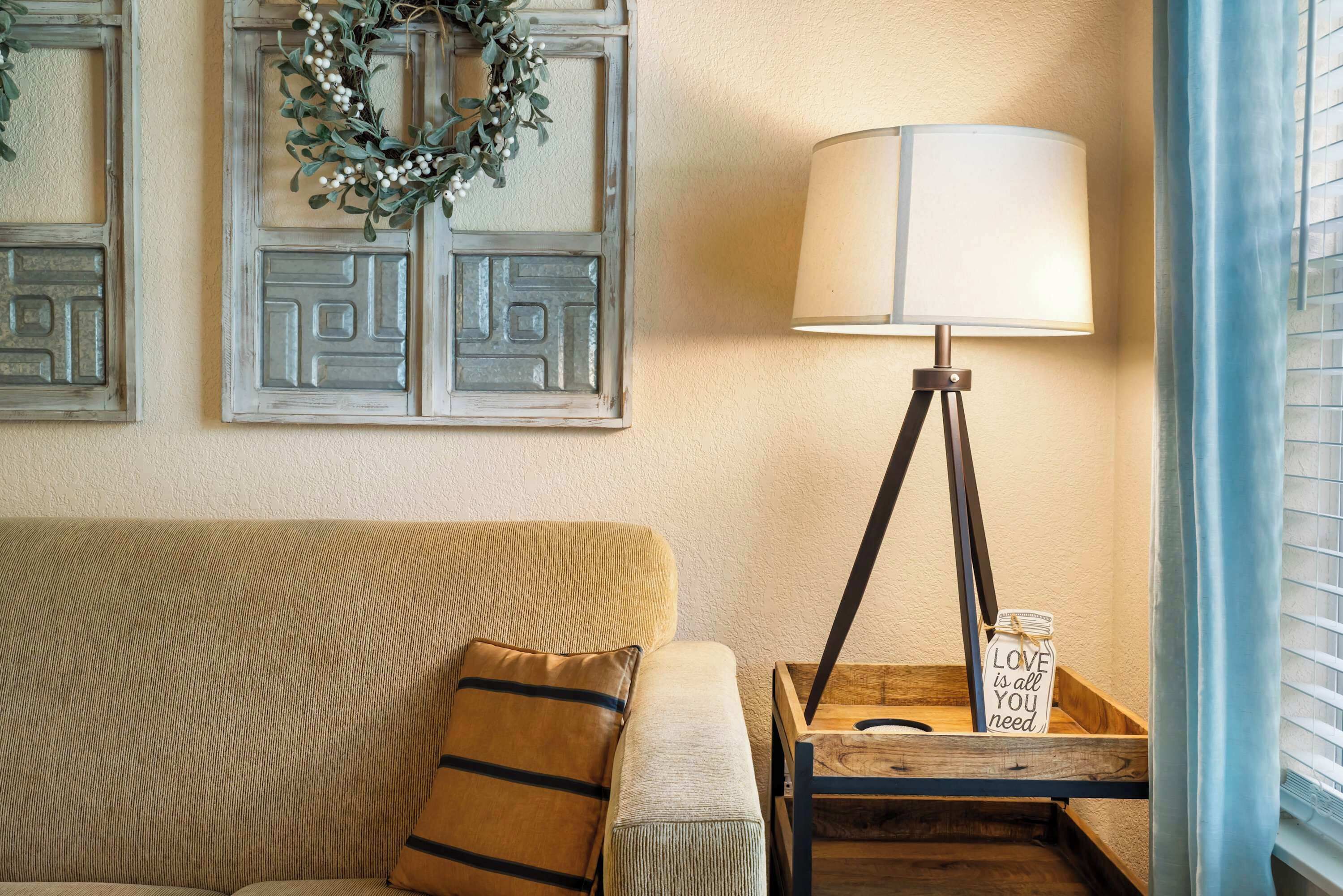 A cozy living room with a beige sofa, a decorative wreath, a lamp on a wooden side table, and a small sign that reads "LOVE is all you need."
