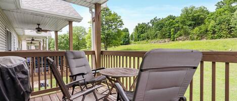 A covered outdoor deck with two chairs and a table, overlooking a grassy area with trees.