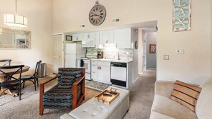 A living room with a sofa, ottoman, and a dining area with chairs and a table. A kitchen is visible in the background with white cabinets and a clock on the wall. - Warm Welcome Getaway Resort Stay for 6 With Amenities Near Downtown (Branson)