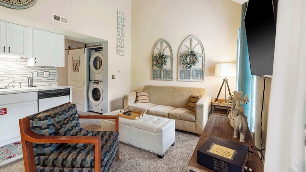 A living room with a sofa, armchair, ottoman, and a small table with a tray. There is a kitchen area with cabinets and a washing machine. - Warm Welcome Getaway Resort Stay for 6 With Amenities Near Downtown (Branson)
