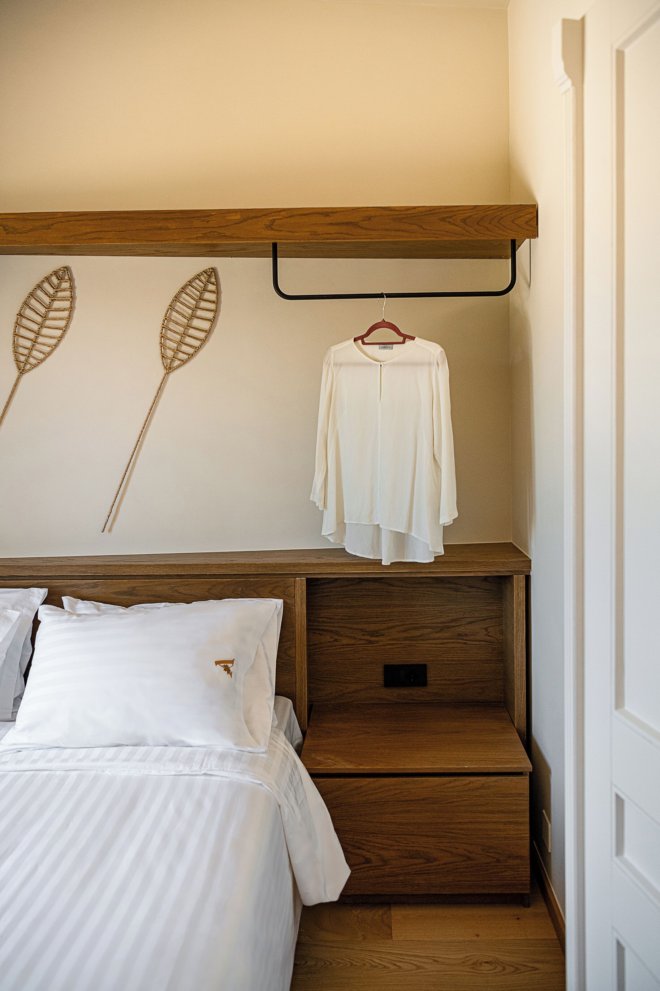 Une chambre à coucher avec une tête de lit en bois, un lit recouvert d’une literie blanche, un vêtement suspendu et deux objets décoratifs fixés au mur.