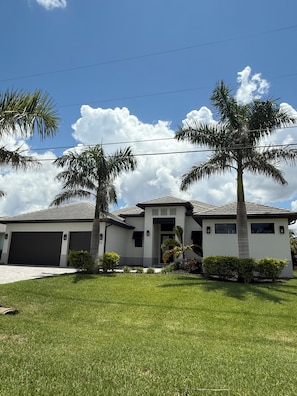 Uma casa moderna de dois andares com garagem, rodeada por gramado bem cuidado e palmeiras.
