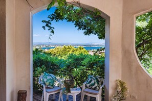 An outdoor seating area with two chairs and a table, overlooking a coastal landscape with trees and a view of the sea.