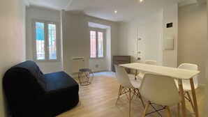 A modern living room with a black sofa, a dining table with white chairs, and large windows with metal bars. - Superb studio with sleeping area! Old Nice! (Nice)