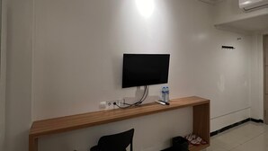 A modern hotel room with a wooden desk, a black chair, a flat-screen TV mounted on the wall, and a small shelf with a water bottle and remote control. - Hotel Harmony (Batulicin)