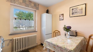 Une cuisine avec une fenêtre, un réfrigérateur, une table recouverte d’une nappe à motifs floraux, un grille-pain et un vase rempli de fleurs.