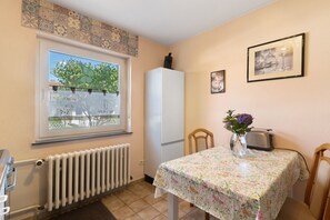 A kitchen with a window, a refrigerator, a table with a floral tablecloth, a toaster, and a vase with flowers.