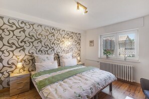 Un dormitorio con papel tapiz estampado, una cama con cubrecama verde, una mesita de noche con lámpara, una ventana con vistas y un radiador.