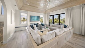 A modern living room with a large L-shaped sectional sofa, a coffee table, and a ceiling fan. There are large windows offering a view of the outdoors.