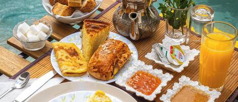 A breakfast setting by a pool with bread, pastries, an omelette, and orange juice.