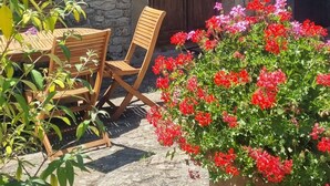 Een stenen gebouw met een houten deur, een grote potplant met rode bloemen en twee houten stoelen.