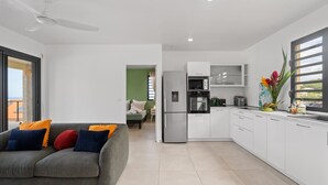Eine moderne Küche mit weißen Schränken, einem eingebauten Backofen und einem Kühlschrank. Im Wohnbereich befindet sich ein graues Sofa mit bunten Kissen.