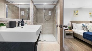 Une salle de bain moderne avec une douche à parois de verre, un meuble-lavabo blanc doté d’un distributeur de savon noir et une table de chevet avec une lampe et une plante.