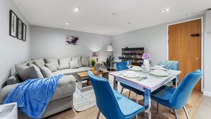 Un salon moderne avec un canapé gris, des fauteuils bleus, une table à manger dressée pour quatre personnes et une bibliothèque en arrière-plan.