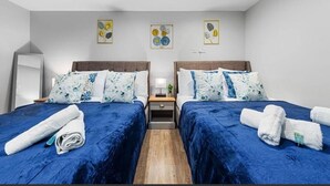 Une chambre double avec deux lits, chacun agrémenté de draps bleus et d’oreillers à motifs floraux. Des appliques murales et des tableaux représentant des motifs floraux encadrés sont disposés au-dessus des lits.