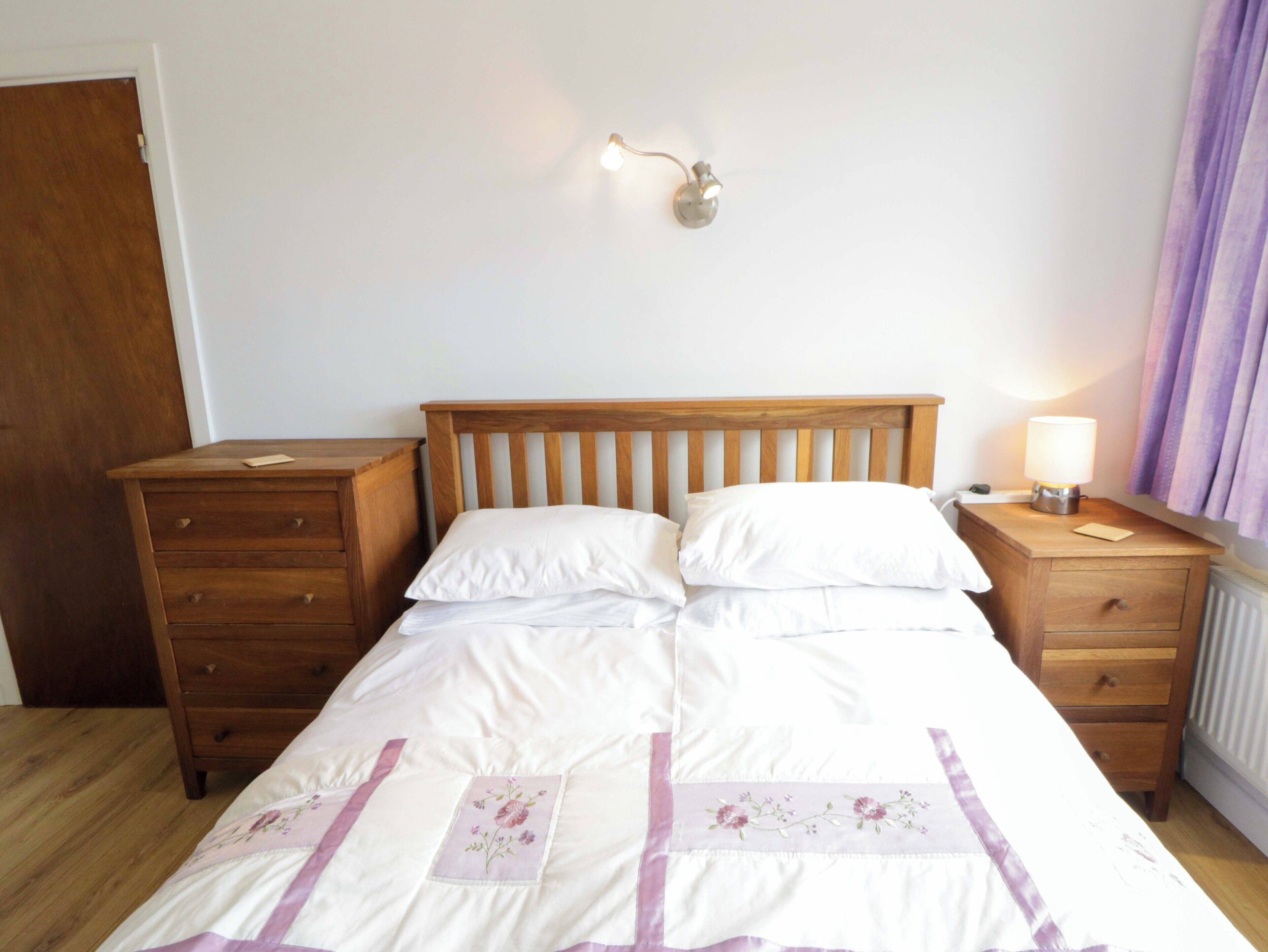 A neatly made bed with a wooden headboard, a bedside table with a lamp, and a wooden dresser.