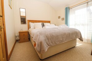 Una cama bien hecha con cabecera de madera, una mesita de noche con lámpara y una ventana con cortinas traslúcidas.