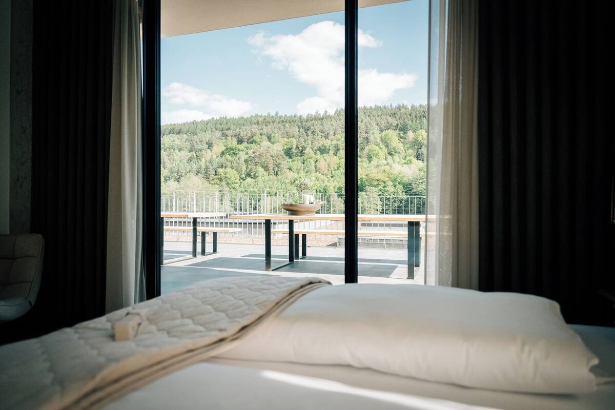A hotel room with a large bed, a view of a forested area, and a balcony with a table and chairs.