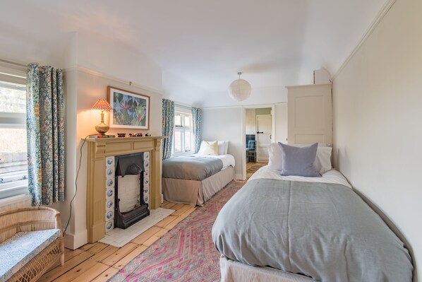 A bedroom with a fireplace, two beds, a wicker chair, and a patterned rug. - Water Farm House, Stowting (Stowting)