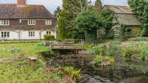 A rustic wooden bridge over a pond, surrounded by lush greenery and a white house with a brown roof. - Water Farm House, Stowting (Stowting)