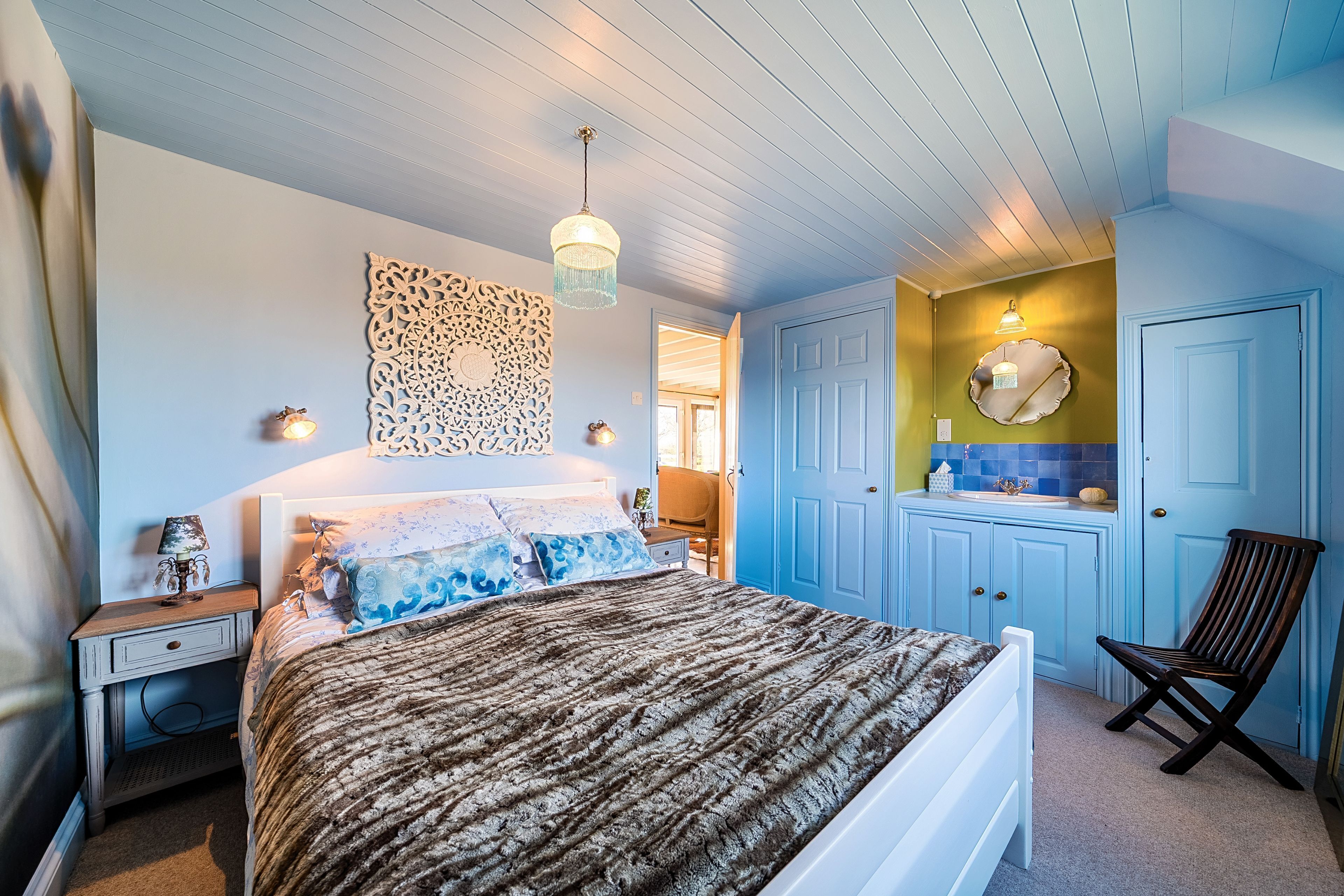 A bedroom with a bed, bedside table, lamp, decorative wall piece, and a chair.