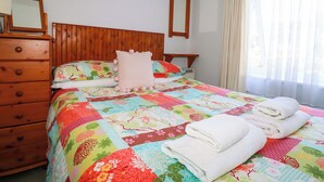 A neatly made bed with a colorful quilt, a wooden dresser, a mirror, and a window with curtains. - Chyancrowse (Penzance)