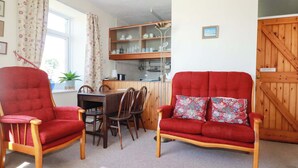 A cozy living room with two red armchairs, a wooden dining table, and a kitchen area with wooden cabinets and a hanging pot rack. - Chyancrowse (Penzance)