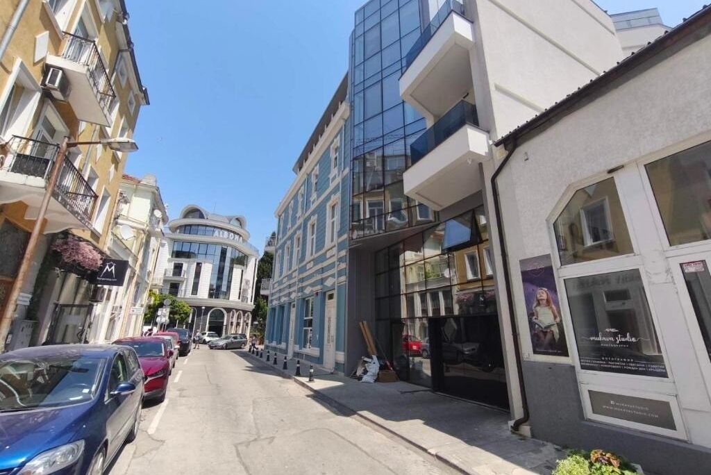 A street with parked cars and buildings, including a modern glass structure and a traditional building with a curved facade.