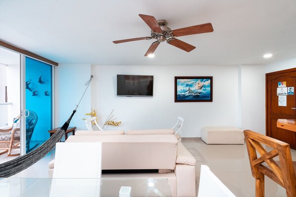 Un salon moderne avec un canapé beige, une table basse en verre, une chaise en bois, un ventilateur de plafond, un téléviseur et un tableau encadré accroché au mur.