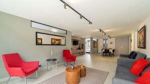 Una sala de estar moderna con una silla roja, sofá gris y mesa de centro de madera. En el fondo, hay un comedor con un televisor y un mueble de madera.