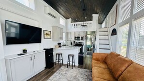 A modern kitchen with white cabinetry, a large TV, and a central island with stools. - The Little Lonestar with Pool & Hot Tub (Austin)