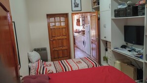 A bedroom with a bed, a desk with a computer, a wardrobe, and a kitchen area visible through an open door. - Nancy and Carlos's house, view of the mountains, swimming pool and barbecue area. (San Luis)