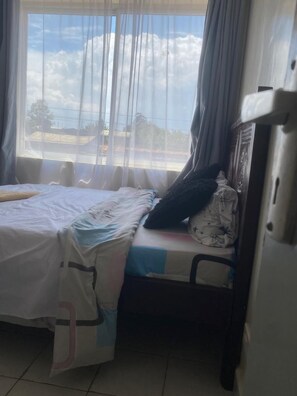 Une pièce avec un lit, une chaise, une fenêtre avec des rideaux et une vue sur le ciel et les nuages.