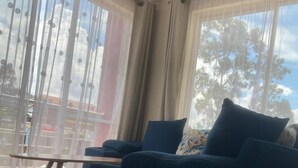 A living room with a dark blue sofa, a small wooden table, and sheer curtains on large windows. - 5 minutes to Ngong hills! Family-sized condo with gym , supermarket $ school (Nairobi)