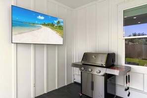 Ein Zimmer mit Flachbildfernseher, der eine Strand Szene zeigt, einem Edelstahlgrill und einem Fenster mit Blick auf einen Garten.