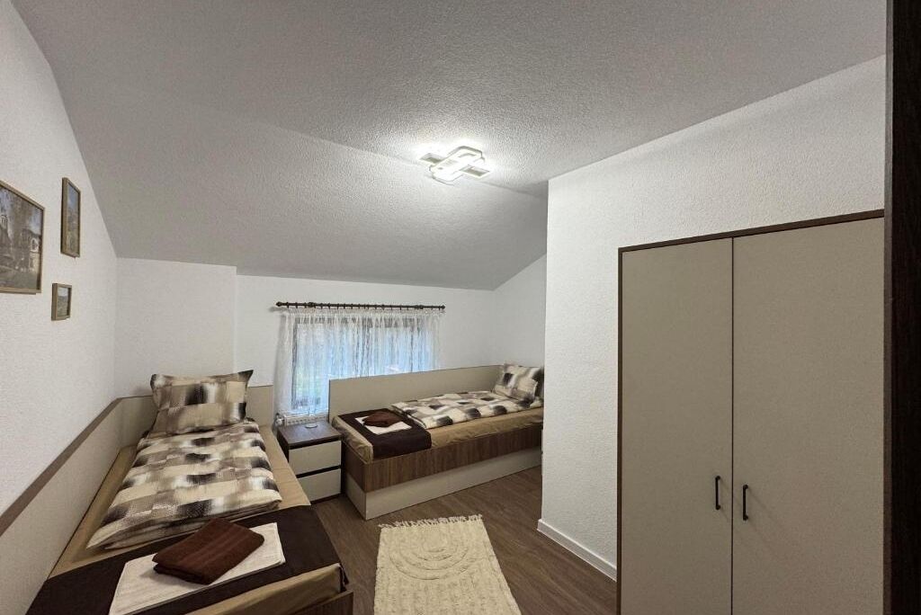 A small, well-lit room with two beds, a wardrobe, and a window with curtains.