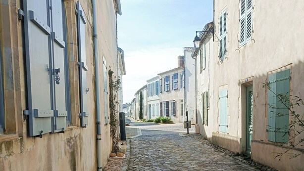 Eine schmale Kopfsteinpflasterstraße, gesäumt von historischen Gebäuden mit verriegelten Fensterläden und einem kleinen Busch.