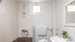 Ein Badezimmer mit weißen Fliesen, einer Duschkabine aus Glas, einer Toilette, einem Waschbecken mit Pflanze und einem Fenster über der Toilette.