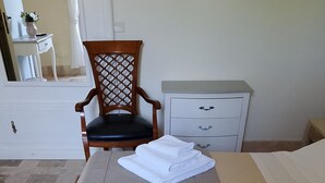 Ein ordentlich bezogenes Bett, gefaltete weiße Handtücher, ein Holzbstuhl, eine weiße Kommode und ein gefliester Boden.