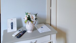 Ein weißes TV-Möbel mit einem flachbildfernseher darüber, einer Vase mit Blumen, einer Fernbedienung und einer Sky-Receiver-Box.