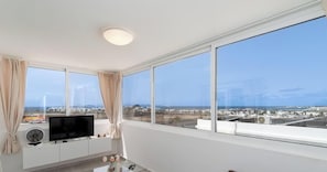 A room with a large window, a flat-screen TV, a small table with a fan, and a view of the cityscape.