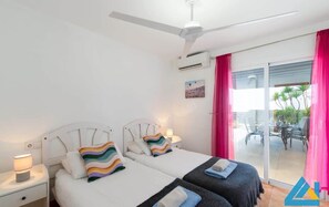 A hotel room with two beds, a ceiling fan, an air conditioning unit, a sliding glass door to the outside, and a small table with a lamp.