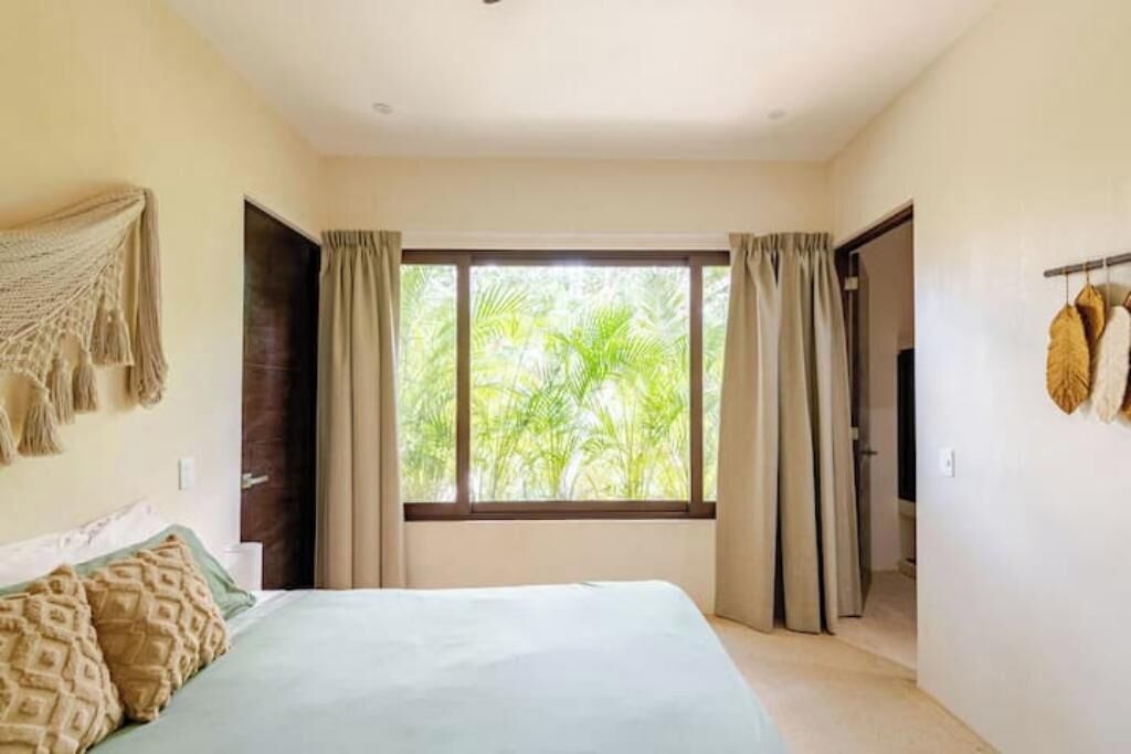 Un dormitorio con cama, una ventana con cortinas, un televisor en la pared y una puerta que da a otra habitación.