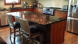 Een keuken met houten kastjes, een granieten eiland, roestvrijstalen apparatuur en een eethoek met rieten stoelen.