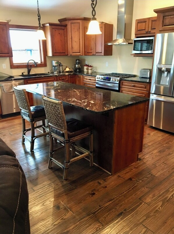 A kitchen with wooden cabinets, a granite island, stainless steel appliances, and a dining area with wicker chairs. - SilverLake House with Hot Tub, game room, huge yard, 1 mile from sand dunes. (Mears)