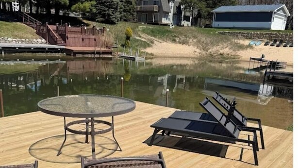 Een houten terras met een glazen tafel en twee lounge fauteuils, met uitzicht op een rustig wateroppervlak en een steiger.